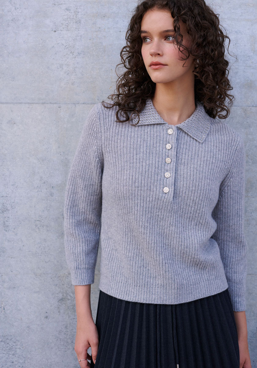 A woman with curly brown hair stands before a light gray concrete wall, wearing a Princess Goes Hollywood ribbed Wolle-Kaschmir-Mix Polokragen sweater with five decorative buttons and a dark pleated skirt. She gazes calmly to her right.