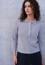 A woman with curly brown hair stands before a light gray concrete wall, wearing a Princess Goes Hollywood ribbed Wolle-Kaschmir-Mix Polokragen sweater with five decorative buttons and a dark pleated skirt. She gazes calmly to her right.