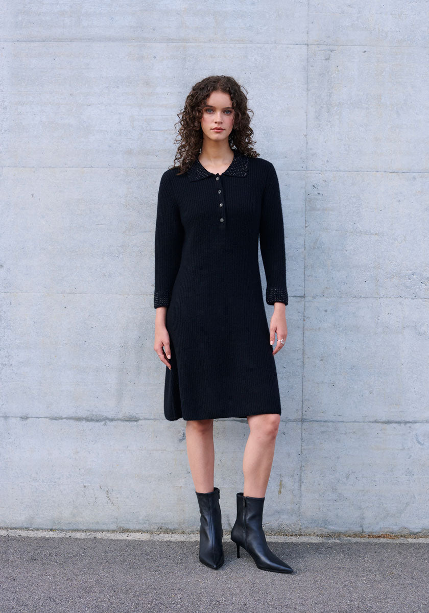 A woman with curly brown hair stands before a light gray concrete wall wearing a long-sleeved, knee-length black wool-cashmere blend polo knit dress with button placket by Princess Goes Hollywood and pointed-toe black ankle boots.