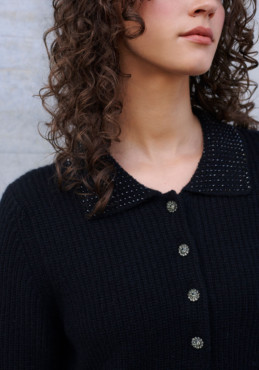 A woman with curly brown hair wears a dark, ribbed knit Princess Goes Hollywood sweater in luxurious wool-cashmere blend with sparkling buttons and a studded polo collar. The image is cropped to her shoulders, her face partially turned away.