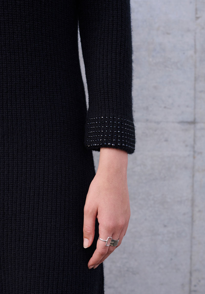 A person’s left arm and hand rest against a gray concrete wall. They wear a Princess Goes Hollywood black polo collar knit dress with small black beads on the cuff and a chunky geometric silver ring on their middle finger.