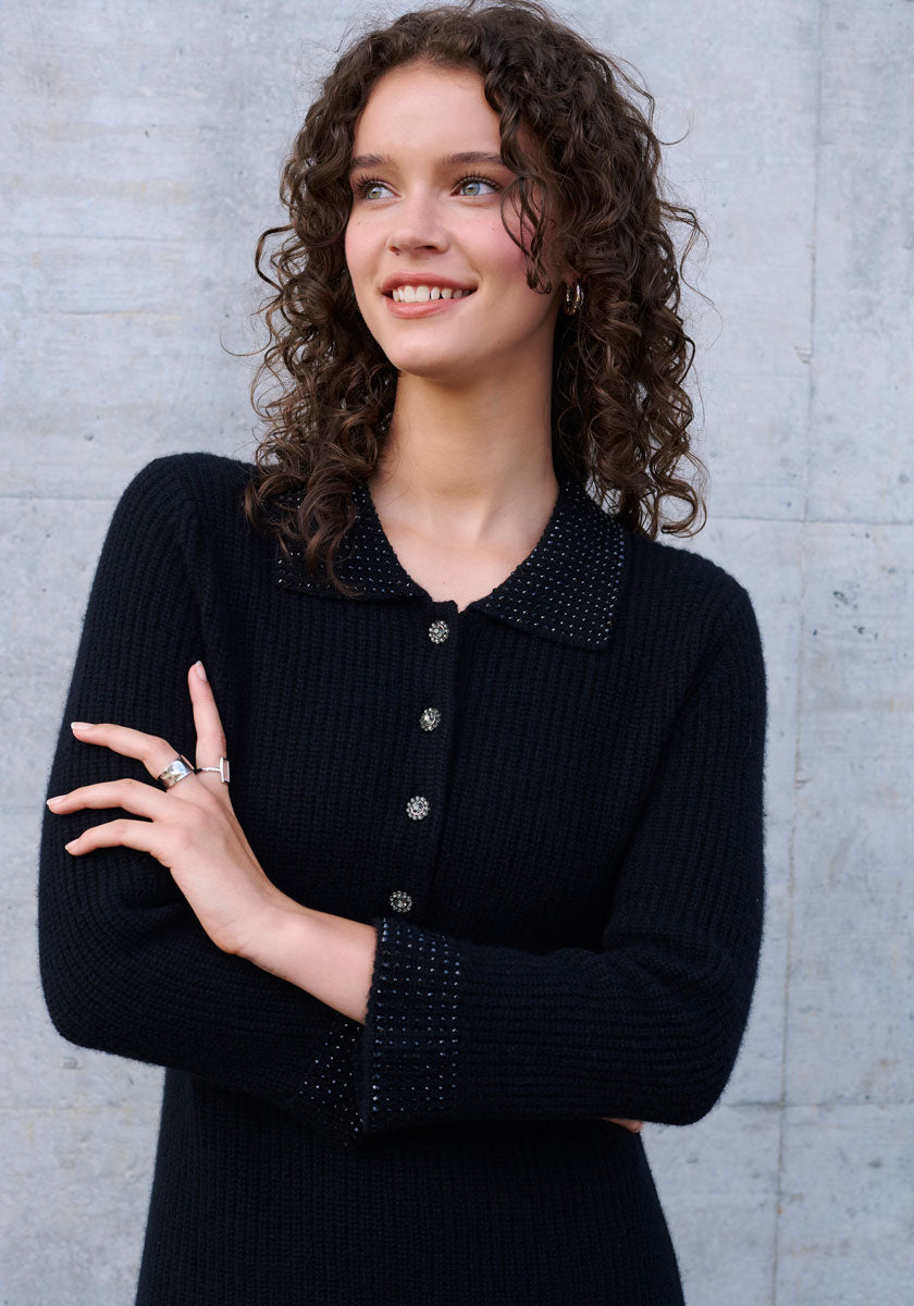 A smiling woman with curly brown hair stands against a light gray textured wall, wearing a Princess Goes Hollywood black wool-cashmere blend knit dress with polo collar, embellished buttons, and pearls on the collar and cuffs. Silver rings adorn her fingers.