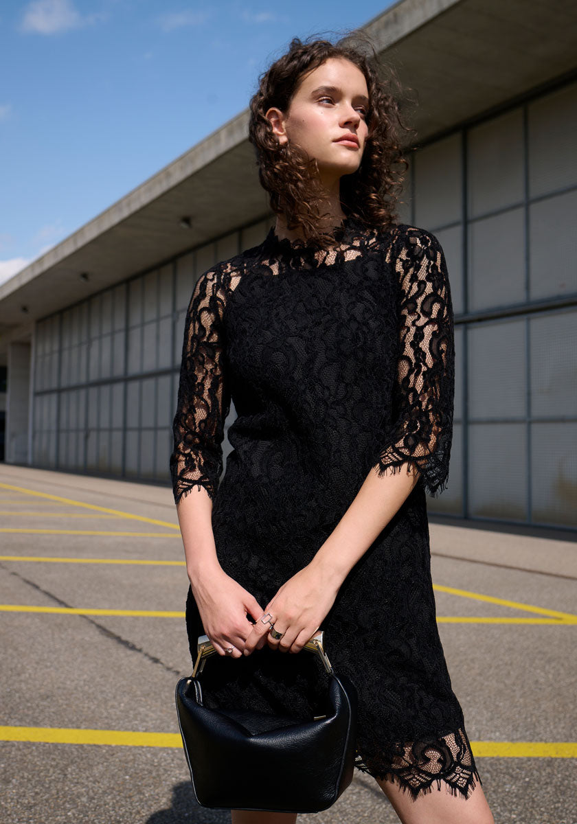 A woman with curly brown hair stands outdoors near a modern building, wearing a Princess Goes Hollywood schwarzes Häkelspitze Kleid mit Stehkragen und 3/4-Ärmeln. She holds a small black handbag with gold handle, looking off to the side as yellow lines mark the ground.