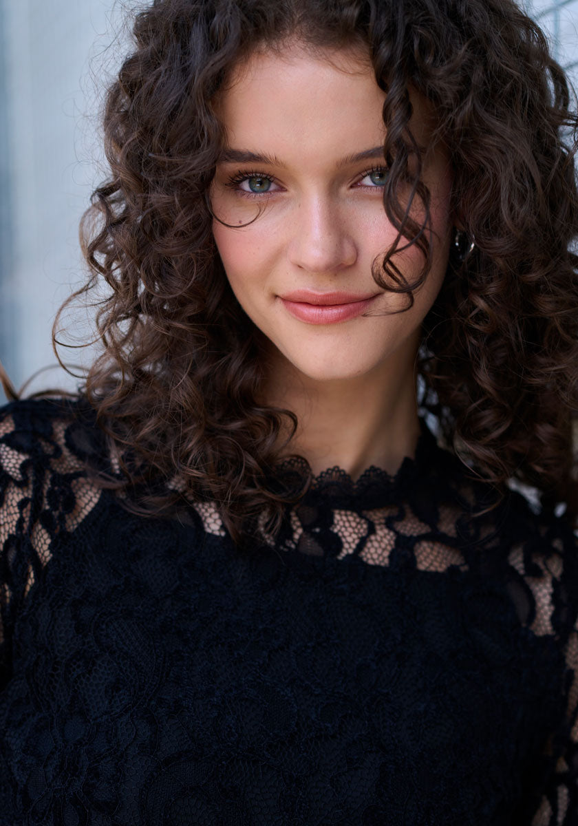 A woman with curly brown hair and blue eyes softly smiles at the camera, wearing a Princess Goes Hollywood Häkelspitze Kleid with transparenten Ärmeln and Stehkragen, standing before a blurred, light-toned background.