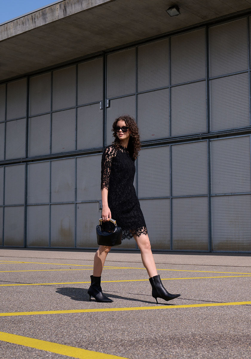 A woman with curly hair and sunglasses strolls through a nearly empty lot, wearing a black Princess Goes Hollywood Häkelspitze Kleid with 3/4-Ärmel, ankle boots, and carrying a chic handbag. An industrial-style metal building stands behind her.