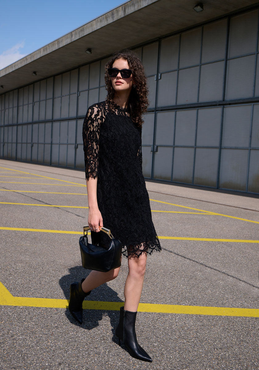 A woman with curly hair in a Princess Goes Hollywood Häkelspitze Kleid with transparent sleeves and sunglasses walks across an empty parking lot, carrying a black handbag. Behind her is a modern building with concrete and metal panels.