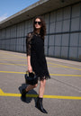 A woman with curly hair in a Princess Goes Hollywood Häkelspitze Kleid with transparent sleeves and sunglasses walks across an empty parking lot, carrying a black handbag. Behind her is a modern building with concrete and metal panels.