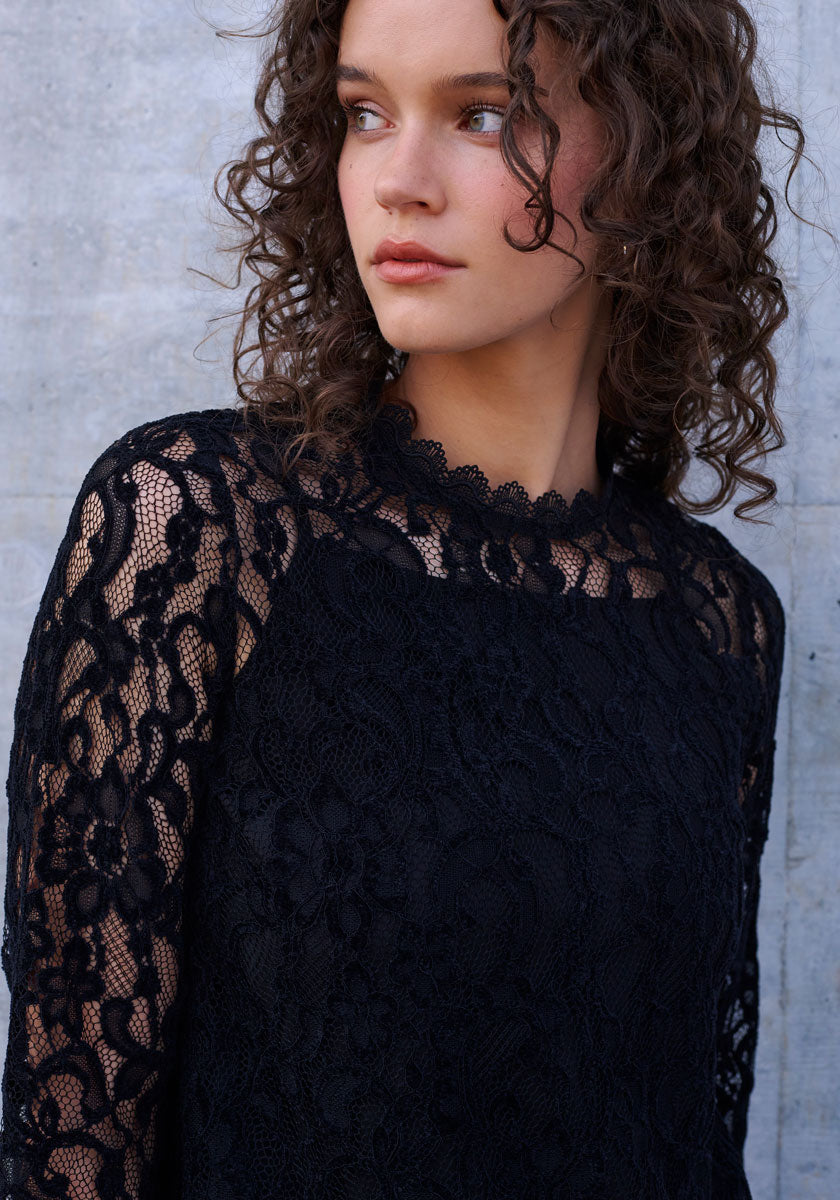 A young woman with curly brown hair gazes left, wearing a Princess Goes Hollywood black lace top with sheer sleeves, floral patterns, and a scalloped neckline. She appears thoughtful against a gray concrete wall.