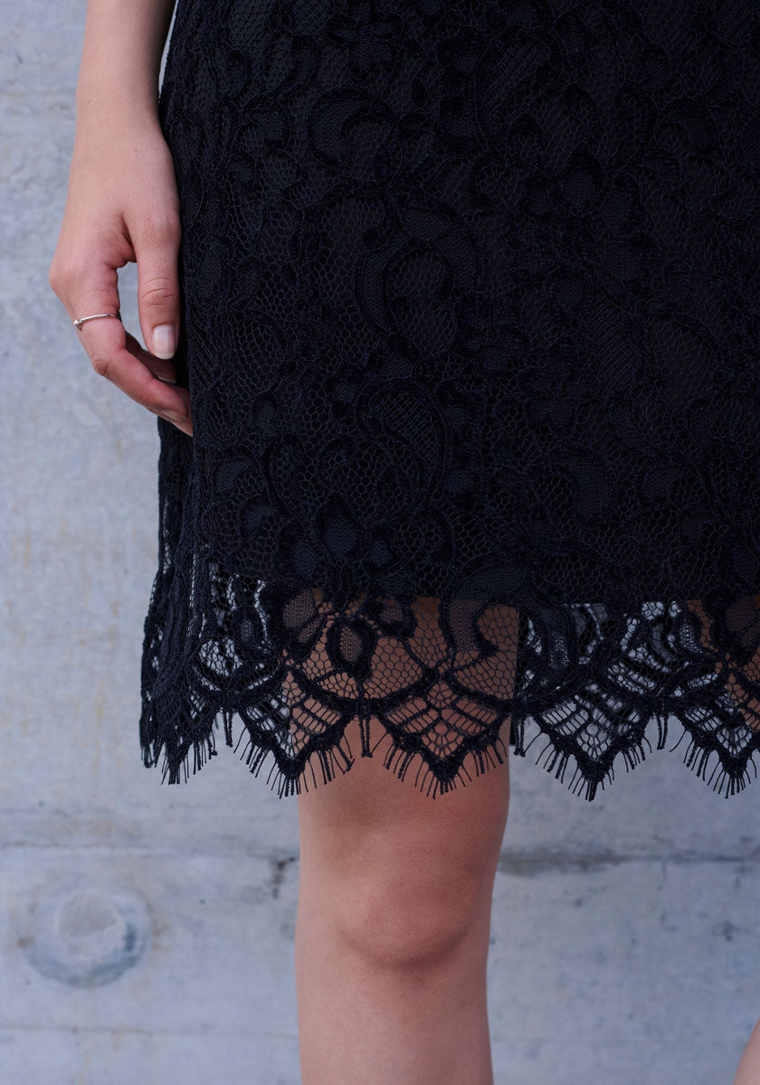 A person stands against a light gray concrete wall, wearing a black Princess Goes Hollywood Häkelspitze Kleid with a scalloped, fringed hem. Their left hand with a thin silver ring rests beside the skirt, revealing part of their bare leg.