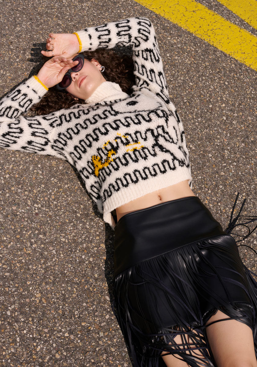 A person with curly hair lies on asphalt, shielding their eyes. They wear Princess Goes Hollywood sunglasses and a cream and black patterned sweater with yellow text, paired with a fringe-detail stylischer Minirock. Midriff is visible.