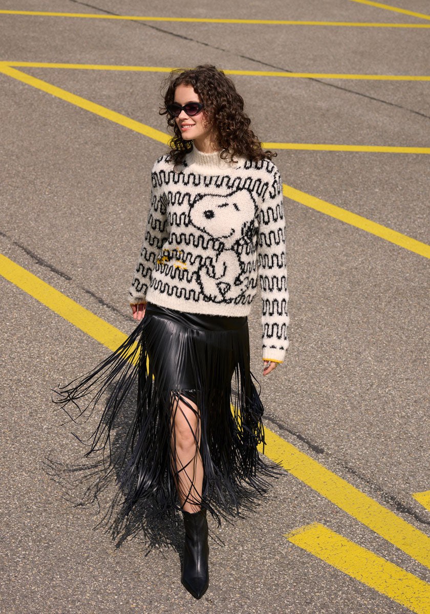 A woman with curly hair and sunglasses wears a cream Princess Goes Hollywood Grobstrickpullover im Wollmix mit Snoopy-Motiv, a black leather skirt with long fringes, and black ankle boots. She smiles while walking on bold yellow lines.
