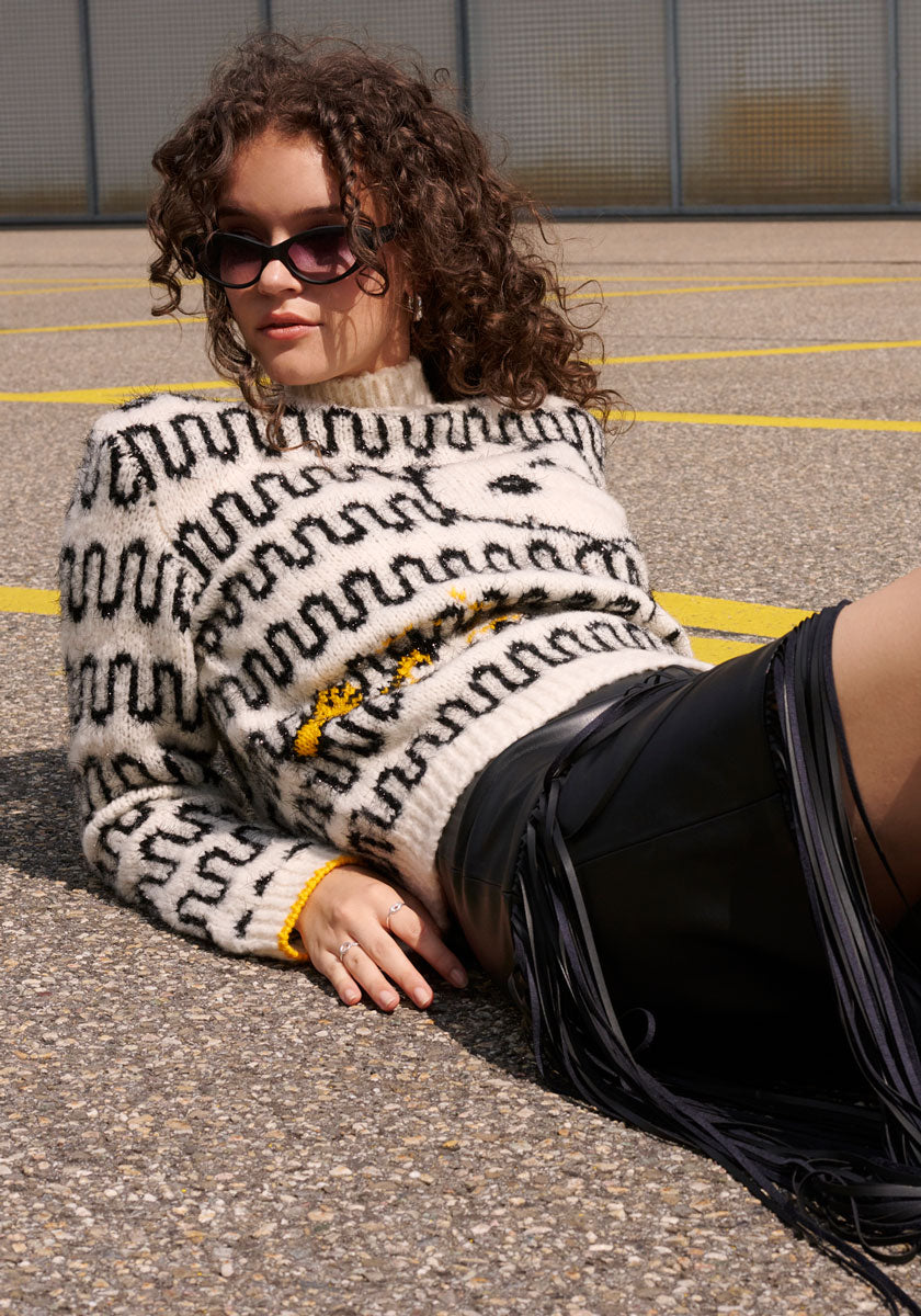 A woman with curly hair in sunglasses lounges on pavement with yellow lines, wearing a Princess Goes Hollywood white Wollmix Grobstrickpullover featuring black squiggly lines and yellow accents, paired with a black fringed skirt. A wire fence is in the background.