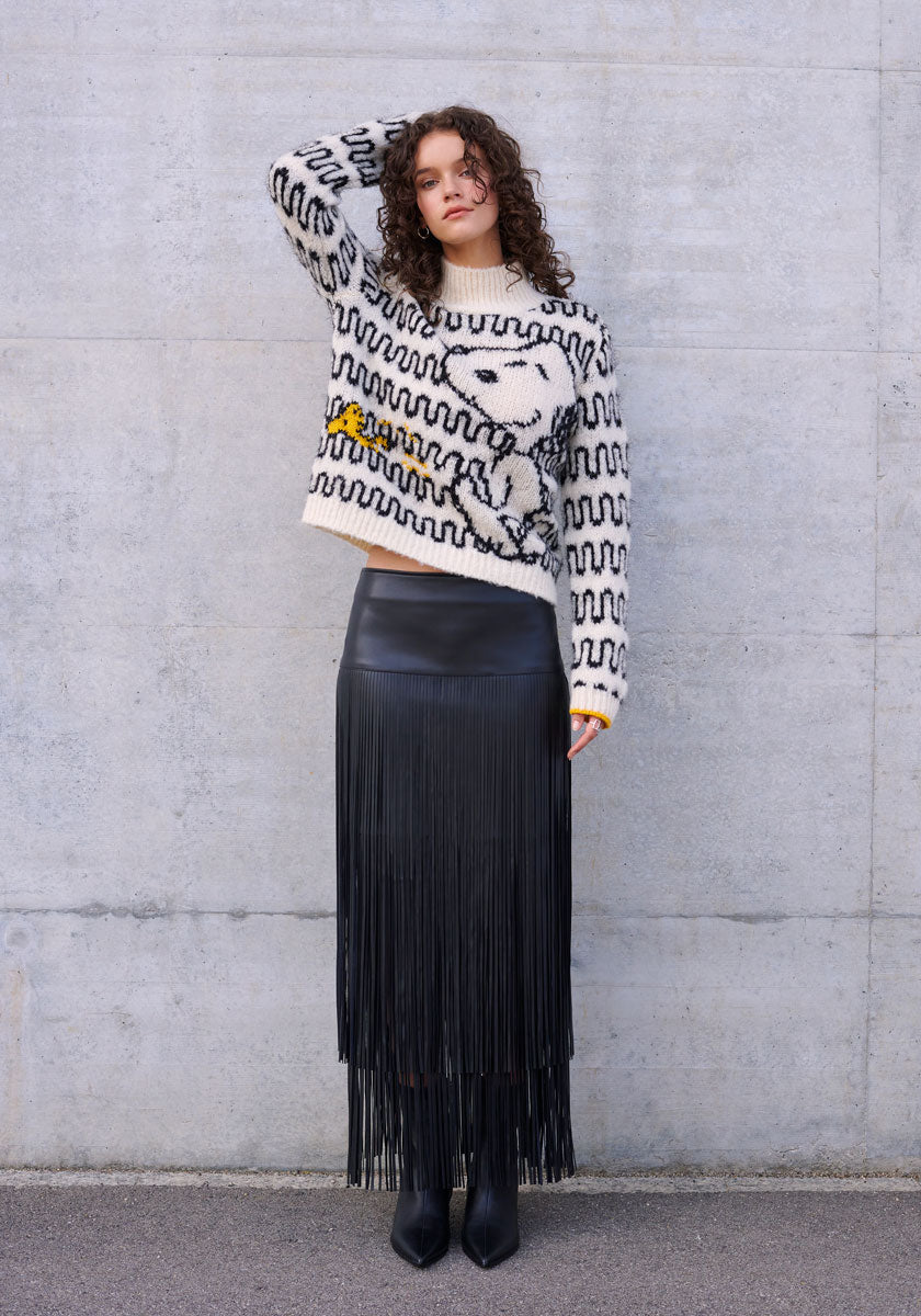 A woman with curly brown hair stands against a gray wall, wearing a fuzzy white Princess Goes Hollywood sweater with a black Snoopy graphic and a stylish faux leather fringe skirt, paired with black ankle boots.