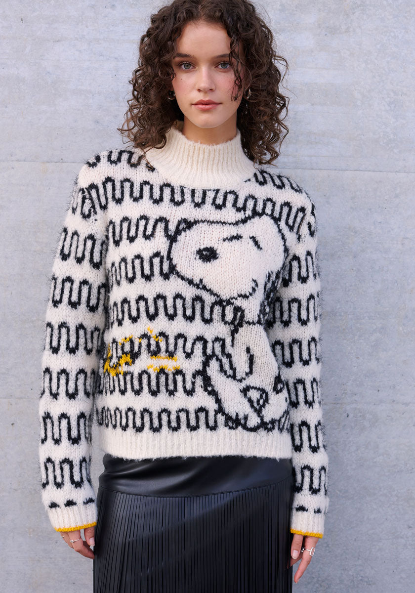 A young woman with curly brown hair wears a Princess Goes Hollywood Wollmix Grobstrickpullover with a large black and white Snoopy and Woodstock motif, paired with a black pleated skirt, standing against a light gray stone wall.