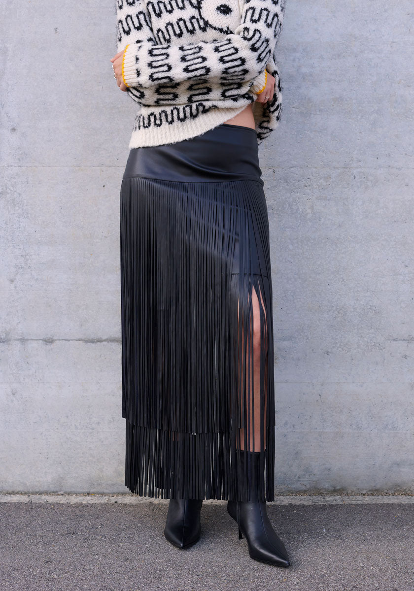 A person stands against a gray wall, wearing a Princess Goes Hollywood black stylischer Kunstleder Minirock mit Fransen, black pointed-toe boots, and a cream sweater with bold black geometric pattern. The photo is cropped above the shoulders.