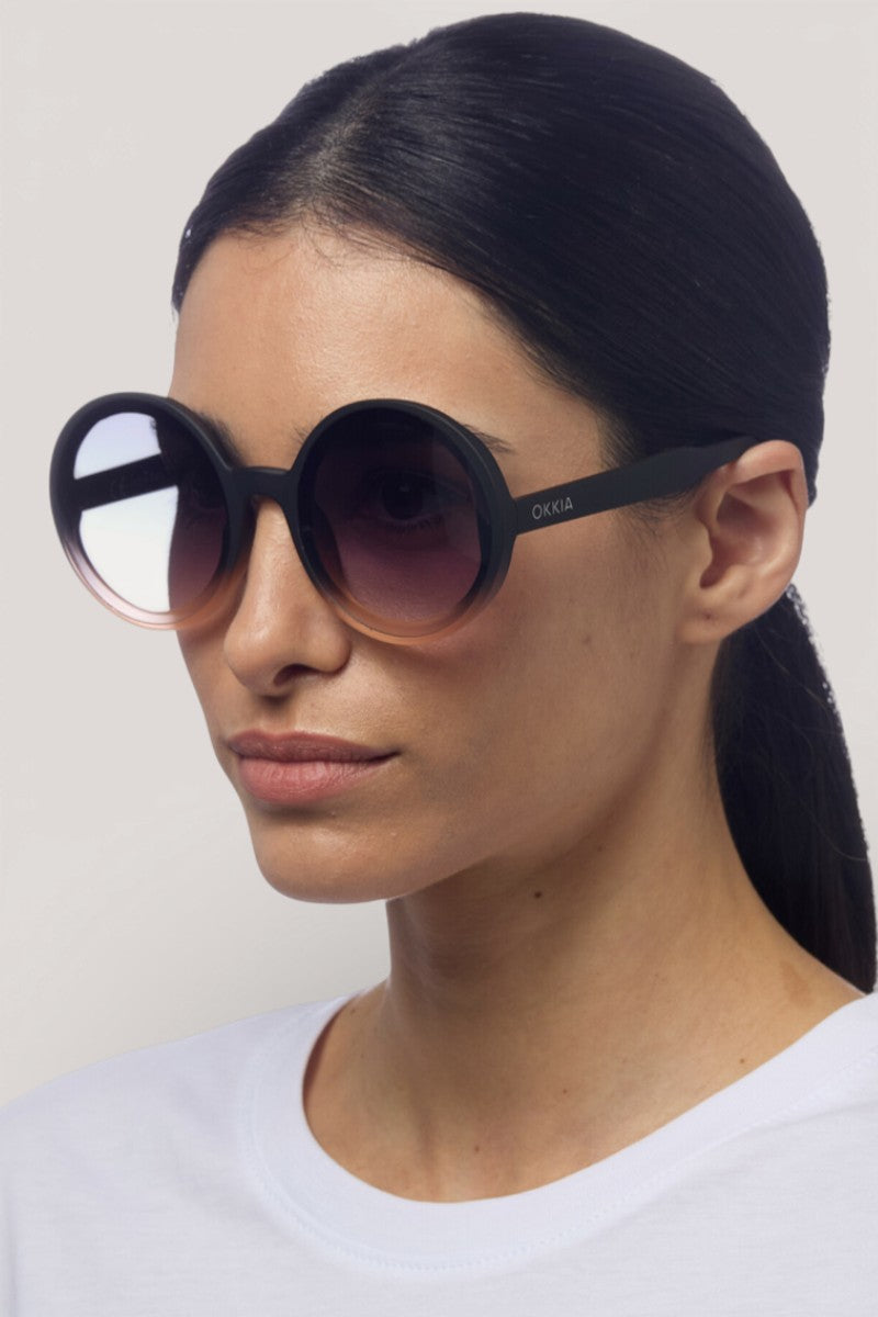 Eine Frau mit langen dunklen Haaren trägt eine runde Sonnenbrille von Okkia in Übergröße mit UV 400 Schutz und Verlaufsgläsern. Sie hat einen neutralen Ausdruck und trägt ein schlichtes weißes Oberteil vor einem hellen Hintergrund.