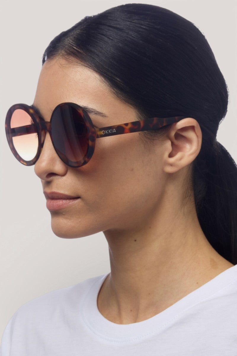Eine Frau mit langen, nach hinten gebundenen dunklen Haaren trägt eine Okkia Oversize Sonnenbrille mit UV 400 Schutz und Verlaufsgläsern, kombiniert mit einem weißen Hemd. Sie ist im Profil mit Blick nach links vor einem schlichten hellgrauen Hintergrund abgebildet.