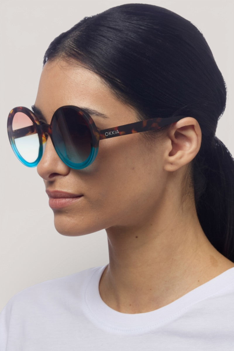 Eine Frau mit glattem, dunklem Haar trägt eine Okkia Oversize Sonnenbrille - eine große, runde Sonnenbrille mit blau getönten Gläsern und schildpattfarbenem Rahmen. Sie schaut leicht nach links in einem weißen Oberteil vor hellem Hintergrund.