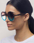 Eine Frau mit glattem, dunklem Haar trägt eine Okkia Oversize Sonnenbrille - eine große, runde Sonnenbrille mit blau getönten Gläsern und schildpattfarbenem Rahmen. Sie schaut leicht nach links in einem weißen Oberteil vor hellem Hintergrund.