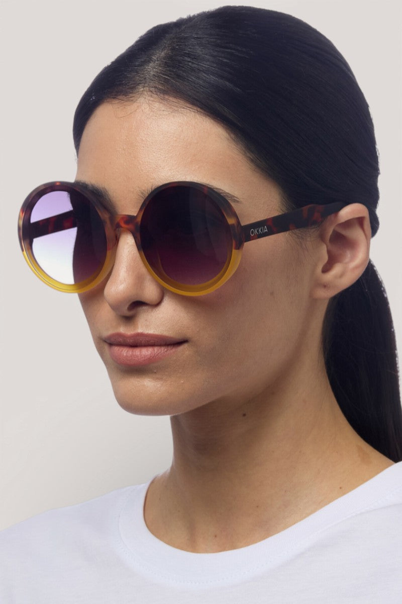 Eine Frau mit glattem, nach hinten gezogenem dunklem Haar trägt eine runde, übergroße Sonnenbrille von Okkia mit UV-400-Faktor und einem Rahmen mit braun-gelbem Farbverlauf. Sie trägt ein einfaches weißes Oberteil und steht vor einem neutralen Hintergrund, wobei sie leicht nach rechts schaut.