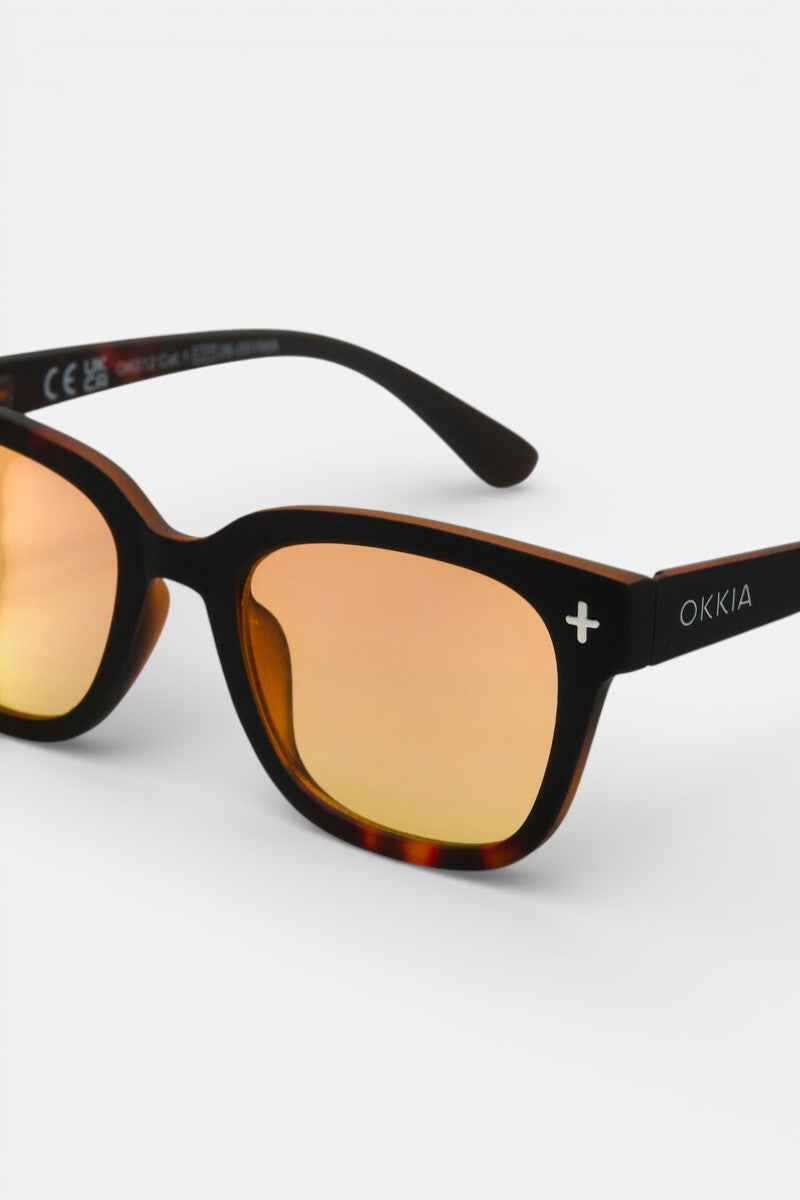 Nahaufnahme der schildpattfarbenen Sonnenbrille von Okkia mit orangefarbenen, verspiegelten Gläsern und quadratischem Design. Der dicke, rechteckige Rahmen ist mit einem weißen Kreuz, dem Okkia-Branding und UV-400-Schutz versehen und steht auf einem schlichten weißen Hintergrund.