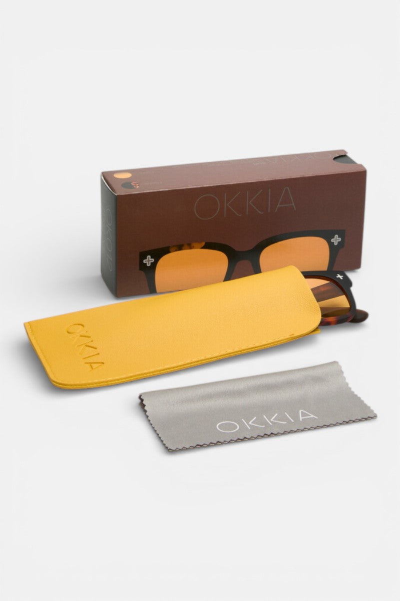 Eine braun gerahmte Okkia-Sonnenbrille mit gelben UV-400-Schutzgläsern sitzt teilweise in einem gelben Etui. Ein graues Putztuch mit der Aufschrift Okkia liegt davor, und eine braune Okkia-Box steht aufrecht vor einem weißen Hintergrund.