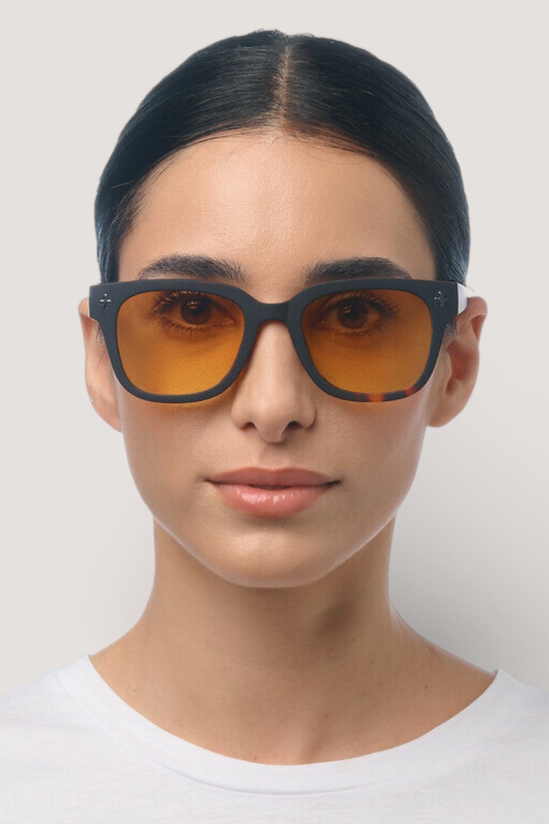 Eine Frau mit hochgestecktem dunklem Haar trägt eine Okkia Sonnenbrille mit quadratischem Rahmen und orangefarbenen UV-400-Schutz-Gläsern sowie ein weißes Hemd. Sie blickt nach vorne vor einem schlichten hellgrauen Hintergrund und behält einen neutralen Ausdruck bei.