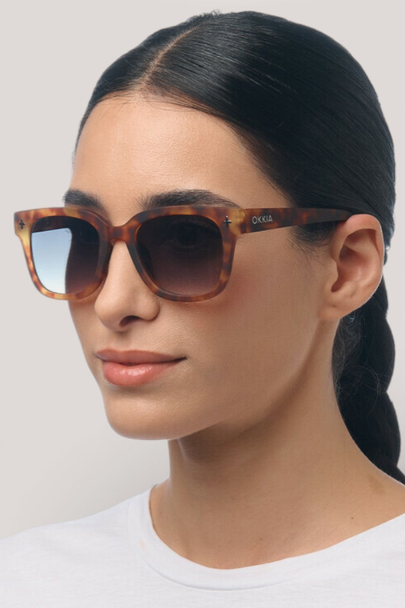 Eine Frau mit glattem schwarzem Haar trägt die Okkia UV 400 Schutz Sonnenbrille mit quadratischem Rahmen und Verlaufsgläsern. Der Markenname "Okkia" ist auf dem Bügel sichtbar. Sie hat einen neutralen Gesichtsausdruck und blickt leicht nach rechts.