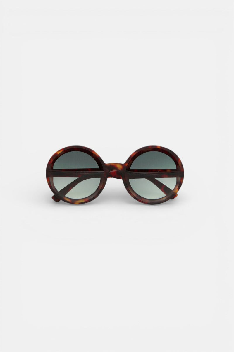 Die runde, übergroße Okkia Damen Sonnenbrille hat einen kräftigen Rahmen aus dunklem Schildpatt, grün getönte Gläser und UV 400-Schutz. Ihr retro-inspiriertes Design wird aufrecht auf einem schlichten weißen Hintergrund dem Betrachter zugewandt präsentiert.