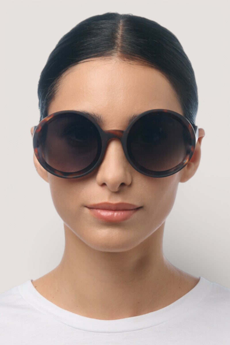 Eine Frau mit mittlerem Hautton und dunklem, zurückgestecktem Haar trägt eine Okkia Oversize Damen Sonnenbrille mit UV 400-Schutz. Sie hat einen neutralen Gesichtsausdruck, ist mit einem schlichten weißen Oberteil bekleidet und steht vor einem hellgrauen Hintergrund.