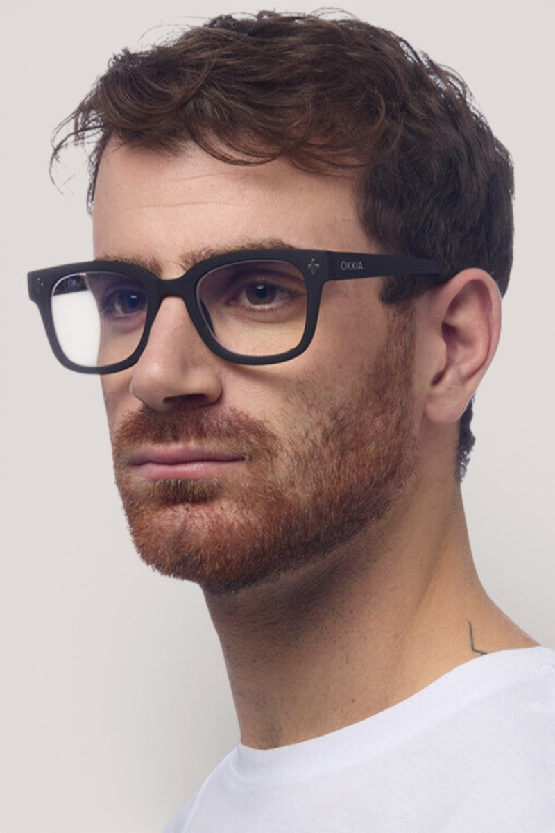 Ein Mann mit kurzen braunen Haaren und einem gestutzten rötlich-braunen Bart trägt eine dicke schwarze Okkia-Bildschirmbrille mit quadratischem Rahmen und ein weißes Hemd. Er blickt leicht nach links vor einem schlichten, hellen Hintergrund, auf dem ein kleines Nackentattoo zu sehen ist.