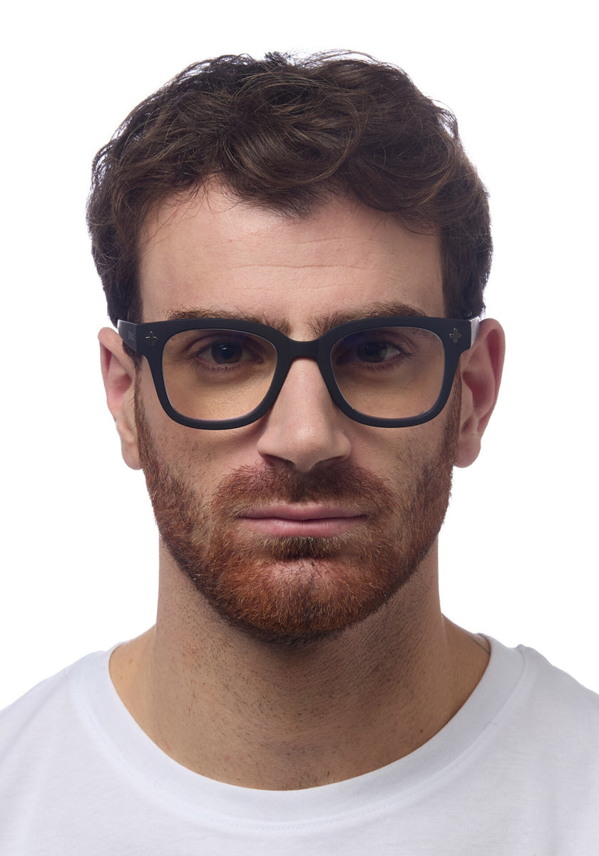 A man with short brown hair and a full reddish-brown beard wears large black Okkia Lesebrille in a soft-touch, square-frame design and a white crew-neck shirt, facing forward with a neutral expression against a plain white background.