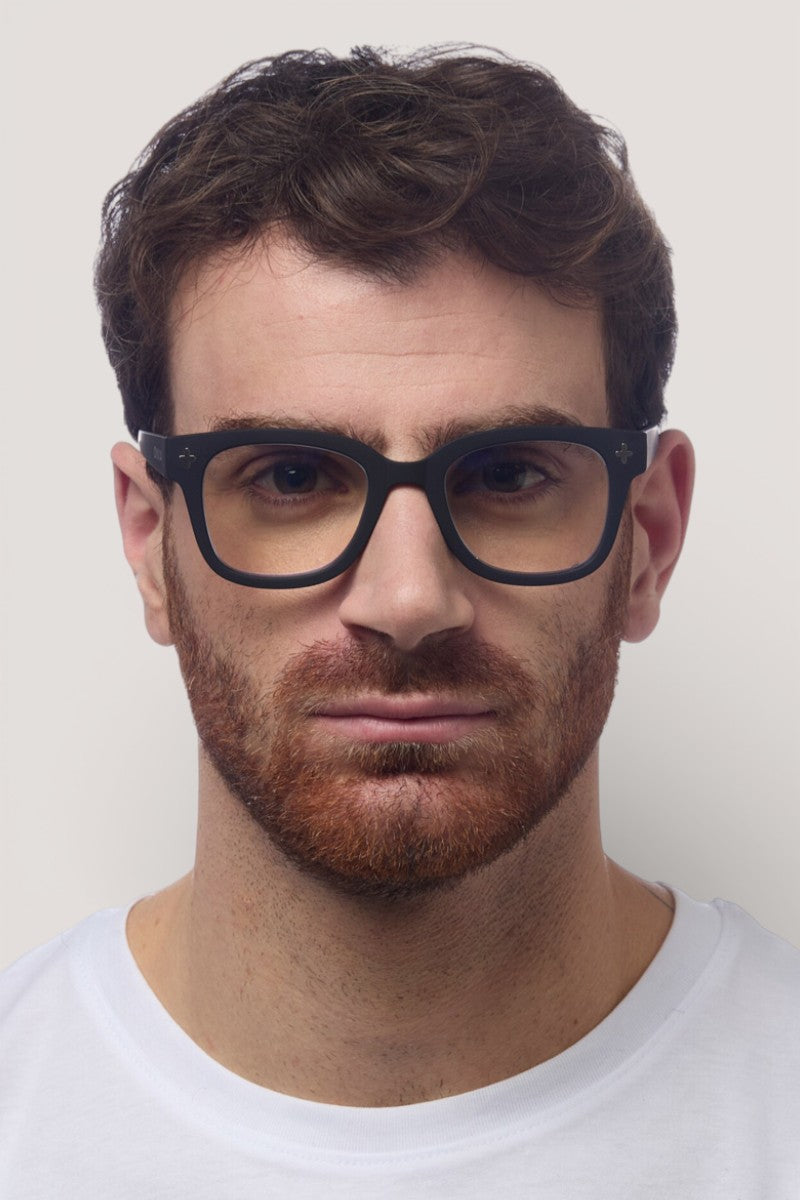 Ein Mann mit kurzen, gewellten braunen Haaren, einem rötlich-braunen Vollbart und einer schwarzen rechteckigen Okkia-Brille mit quadratischem Rahmen trägt ein schlichtes weißes Hemd mit Rundhalsausschnitt und blickt direkt in die Kamera vor einem hellgrauen Hintergrund.