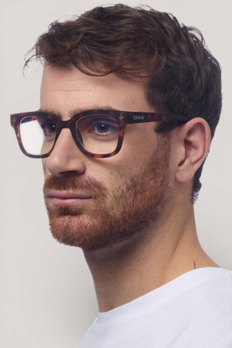 Ein Mann mit kurzen braunen Haaren und einem gestutzten rötlichen Bart trägt ein weißes Hemd und Okkia Square-Frame-Design Blaulichtfilterbrille. Er blickt mit neutraler Miene nach links, seine Brille sorgt für Tragekomfort vor einem schlichten hellen Hintergrund.