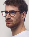 Ein Mann mit kurzen braunen Haaren und einem gestutzten rötlichen Bart trägt ein weißes Hemd und Okkia Square-Frame-Design Blaulichtfilterbrille. Er blickt mit neutraler Miene nach links, seine Brille sorgt für Tragekomfort vor einem schlichten hellen Hintergrund.
