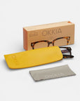 Eine quadratische Schildpattbrille von Okkia in einem gelben Etui, begleitet von einem grauen Putztuch und einer hellbraunen OKKIA-Schachtel mit einer Brillenillendarstellung auf weißem Hintergrund.