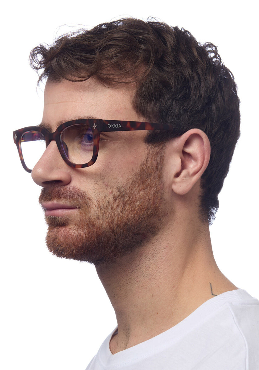A man with short, wavy brown hair and a trimmed beard is shown in profile, wearing Okkia square-frame Lesebrille with a soft-touch frame. He pairs them with a plain white T-shirt against a white background.