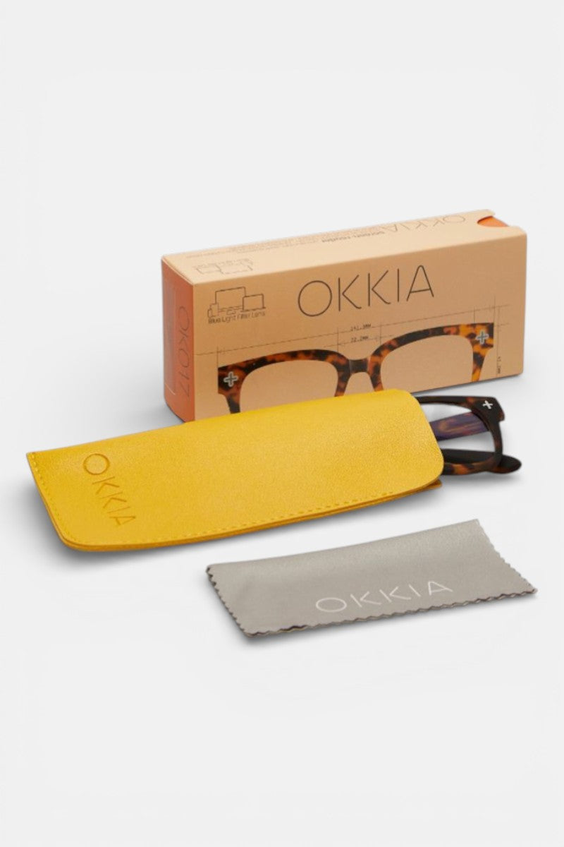 Okkia-Schildpatt-Lesebrille mit quadratischem Rahmen, teilweise in einem gelben Etui, darunter ein graues Putztuch, ausgestellt vor einer beigen Okkia-Schachtel mit Brillengrafik. Alles vor einem schlichten weißen Hintergrund.