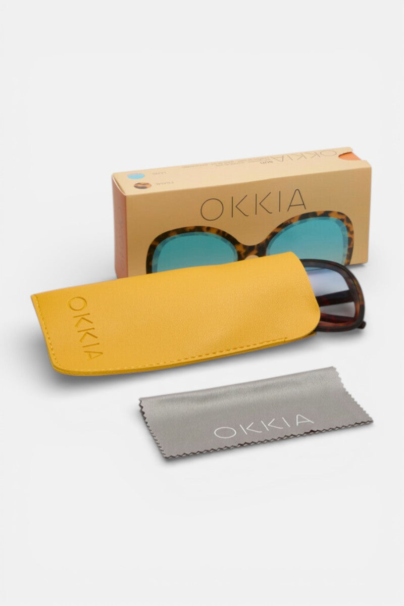 Eine Sonnenbrille von Okkia im Schmetterlingsdesign aus Schildpatt mit blauen UV400-Gläsern, teilweise in einem gelben Okkia-Etui, dazu ein graues Reinigungstuch und eine braune Schachtel mit Sichtfenster, alles auf weißem Hintergrund.