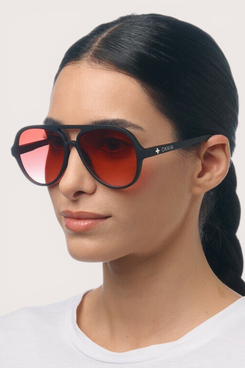 Eine Frau mit langen, nach hinten gebundenen dunklen Haaren trägt ein weißes Hemd und eine elegante Okkia Aviator-Sonnenbrille mit schwarzem Rahmen und großen, rot getönten Gläsern. Sie blickt mit ruhigem Gesichtsausdruck leicht nach rechts. Der Markenname „Okkia“ ist auf dem Bügel der Brille zu sehen und unterstreicht deren Unisex-Design sowie den zuverlässigen UV-400-Schutz für den täglichen Gebrauch.