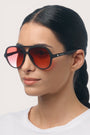Eine Frau mit langen, nach hinten gebundenen dunklen Haaren trägt ein weißes Hemd und eine elegante Okkia Aviator-Sonnenbrille mit schwarzem Rahmen und großen, rot getönten Gläsern. Sie blickt mit ruhigem Gesichtsausdruck leicht nach rechts. Der Markenname „Okkia“ ist auf dem Bügel der Brille zu sehen und unterstreicht deren Unisex-Design sowie den zuverlässigen UV-400-Schutz für den täglichen Gebrauch.