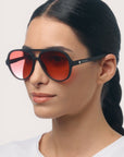 Eine Frau mit langen, nach hinten gebundenen dunklen Haaren trägt ein weißes Hemd und eine elegante Okkia Aviator-Sonnenbrille mit schwarzem Rahmen und großen, rot getönten Gläsern. Sie blickt mit ruhigem Gesichtsausdruck leicht nach rechts. Der Markenname „Okkia“ ist auf dem Bügel der Brille zu sehen und unterstreicht deren Unisex-Design sowie den zuverlässigen UV-400-Schutz für den täglichen Gebrauch.