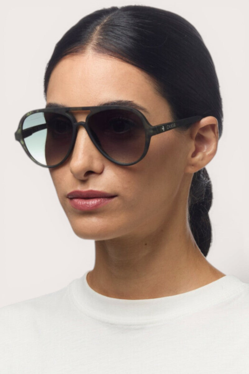 Eine Frau mit dunklem, nach hinten gezogenem Haar trägt eine Okkia Aviator Sonnenbrille mit UV 400 Schutz und ein weißes Oberteil mit Rundhalsausschnitt. Sie blickt leicht nach links vor einem schlichten, hellen Hintergrund und behält einen neutralen Ausdruck bei.