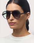Eine Frau mit dunklem, nach hinten gezogenem Haar trägt eine Okkia Aviator Sonnenbrille mit UV 400 Schutz und ein weißes Oberteil mit Rundhalsausschnitt. Sie blickt leicht nach links vor einem schlichten, hellen Hintergrund und behält einen neutralen Ausdruck bei.