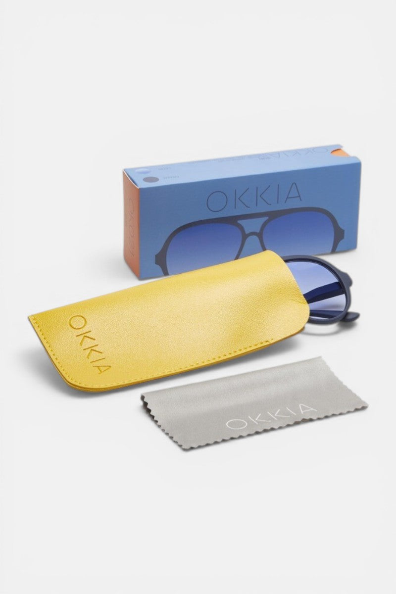 Eine Okkia Aviator-Sonnenbrille mit blauen Gläsern und UV-400-Schutz lugt aus einem gelben Okkia-Etui hervor. Ein graues Okkia-Putztuch und eine bunte blau-orangefarbene Box mit dem Markennamen vervollständigen dieses Unisex-Set.