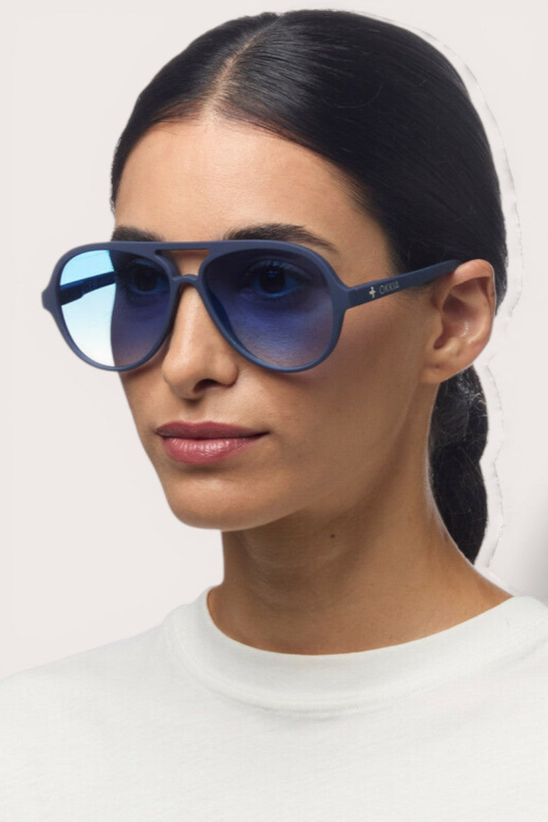 Eine Frau mit glattem, dunklem Haar trägt eine reflektierende blaue Okkia Aviator Sonnenbrille mit UV 400 Schutz und ein weißes Hemd. Sie hat einen neutralen Gesichtsausdruck und steht vor einem schlichten, hellen Hintergrund.