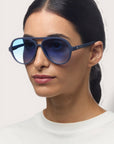 Eine Frau mit glattem, dunklem Haar trägt eine reflektierende blaue Okkia Aviator Sonnenbrille mit UV 400 Schutz und ein weißes Hemd. Sie hat einen neutralen Gesichtsausdruck und steht vor einem schlichten, hellen Hintergrund.