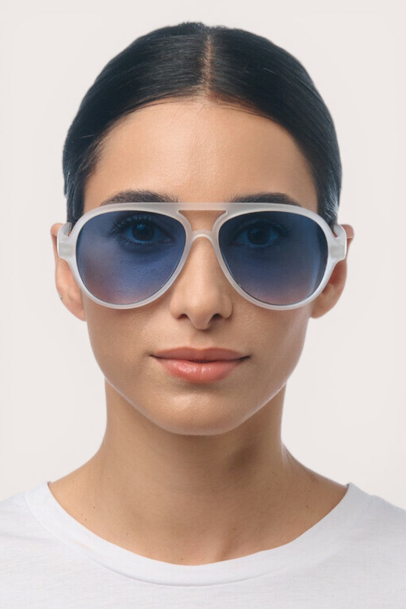 Eine Person mit glattem, dunklem Haar trägt eine große Okkia Aviator-Sonnenbrille mit UV-400-Schutz, weißem Rahmen und blau getönten Gläsern. Sie hat einen neutralen Gesichtsausdruck und trägt ein schlichtes weißes Hemd vor einem hellen Hintergrund.