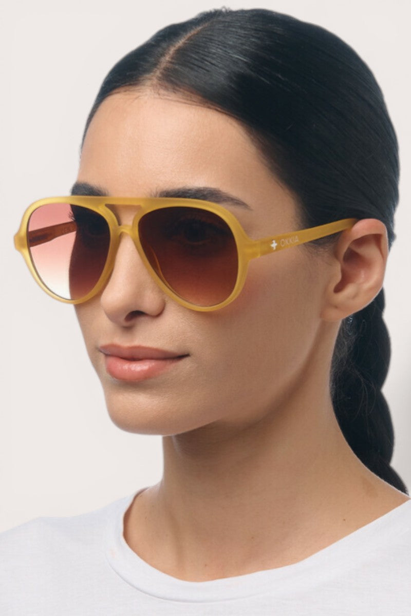 Eine Frau mit glattem, zurückgebundenem dunklem Haar trägt eine gelbe Okkia Aviator-Sonnenbrille mit UV-400-Schutz und braunen Gläsern. Sie hat einen neutralen Gesichtsausdruck und trägt ein schlichtes weißes Oberteil vor einem hellen Hintergrund.