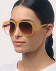 Eine Frau mit glattem, zurückgebundenem dunklem Haar trägt eine gelbe Okkia Aviator-Sonnenbrille mit UV-400-Schutz und braunen Gläsern. Sie hat einen neutralen Gesichtsausdruck und trägt ein schlichtes weißes Oberteil vor einem hellen Hintergrund.